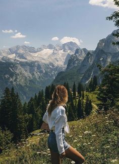 a woman standing on top of a lush green hillside next to mountains with snow capped peaks in the distance