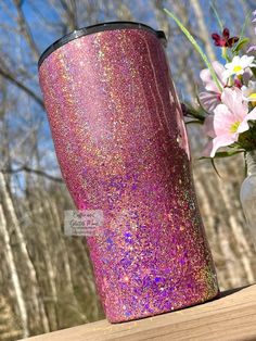 a pink and purple glitter tumbler next to flowers on a wooden table with trees in the background