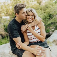 a young man and woman sitting on top of a rock hugging each other with trees in the background