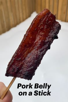 a hand holding a stick with pork belly on a stick in the foreground and text pork belly on a stick above it