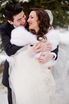 a bride and groom are hugging in the snow with their fur stole around their shoulders