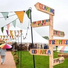 a person with an umbrella walks down a path lined with wooden signs that read, glamping terrace, firepits, kids area, and tents