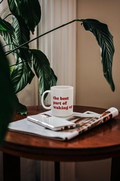 a white coffee mug sitting on top of a wooden table next to a green plant