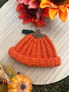 an orange knitted pumpkin hat sitting on top of a wooden board next to flowers