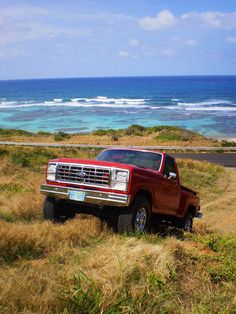 a red pick up truck parked on top of a grass covered hill next to the ocean