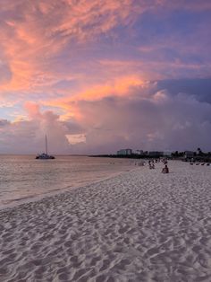 people walking on the beach at sunset with boats in the water and pink clouds above