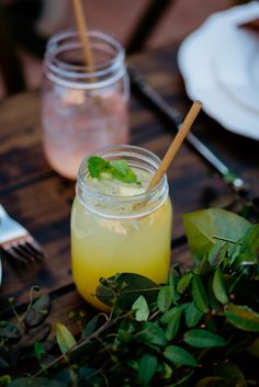 a table topped with two mason jars filled with yellow liquid and green leaves next to utensils