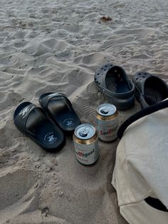 two pairs of slippers sitting on top of a sandy beach next to cans of beer