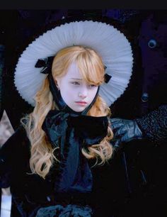 a woman with long blonde hair wearing a white hat and black dress, standing in front of a blue background