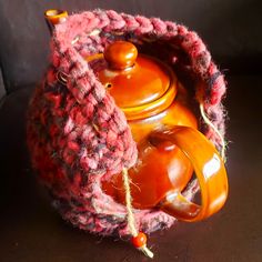 an orange tea pot in a crocheted basket on a brown surface with red yarn