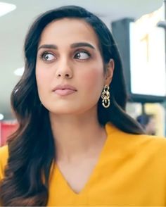 a woman with long black hair wearing gold earrings and a yellow top looking up at the camera