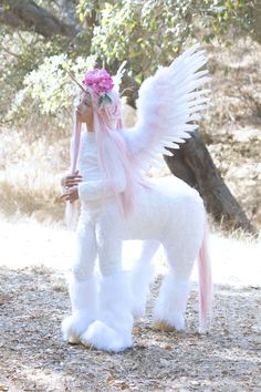 a white dog with wings and flowers on it's head standing in the dirt