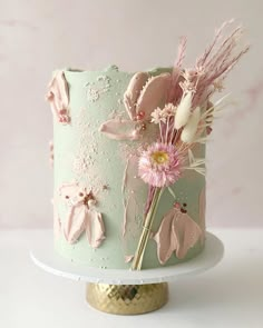 a cake decorated with pink flowers and feathers on a gold plated stand against a pale background