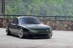 a black car parked in front of a stone wall next to a parking lot with trees behind it