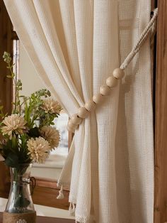 a vase filled with flowers sitting next to a window covered in white sheer curtains and tassels