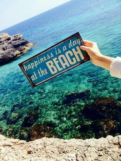 someone holding up a sign that says happiness is a day at the beach in front of some water