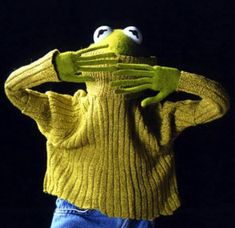 the frog is wearing a yellow sweater and holding his hands up to his face while standing in front of a black background