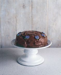 a cake sitting on top of a white cake plate covered in chocolate frosting and blue flowers