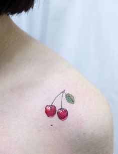 a small cherry tattoo on the back of a woman's left shoulder, with two cherries attached to it