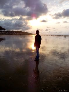 a person standing in the water on a beach at sunset with clouds and sun behind them