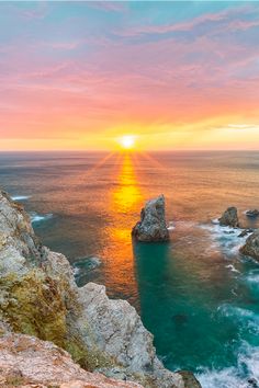 the sun is setting over the ocean with rocks and cliffs in the foreground,