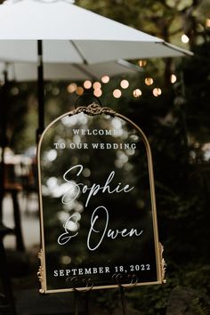 a welcome sign for a wedding with lights in the background and an umbrella over it