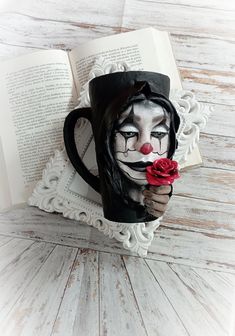 a coffee mug with a clown face holding a rose in it's mouth, sitting on top of an open book