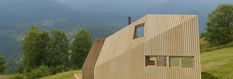 an unusual wooden house on top of a hill with mountains in the background and trees around it