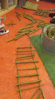 two children are sitting on the floor and one child is standing in front of a bucket full of sticks