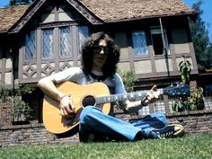 a man sitting on the grass playing a guitar in front of a house with windows