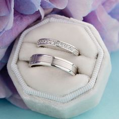 two wedding rings sitting in a white box with purple flowers behind them on a blue background