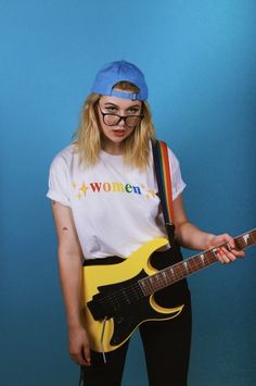 a woman with glasses holding a guitar in front of a blue background and posing for the camera