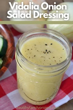 a mason jar filled with salad dressing next to cucumbers and onions on a checkered tablecloth