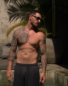 a shirtless man with tattoos standing in front of a palm tree and looking at the camera