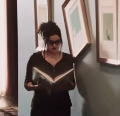 a woman reading a book while standing next to a wall with framed pictures on it