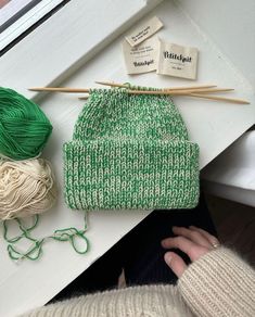 a green hat sitting on top of a white table next to yarn and knitting needles