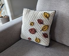 a crocheted pillow with autumn leaves on it sitting on a gray couch in a living room