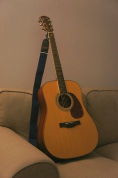 an acoustic guitar sitting on top of a white couch next to a blue strap that is attached to it