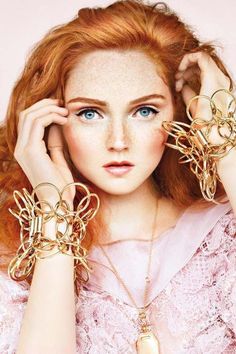 a woman with red hair and blue eyes wearing gold bracelets, holding her hands up to her face