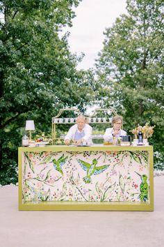 two people sitting at a table in front of some trees and bushes with flowers on it