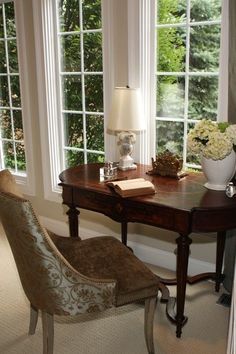 a desk with a lamp and chair in front of two windows that look out onto the trees