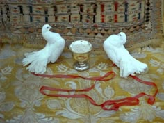 two white doves sitting on top of a table next to a silver cup and red ribbon