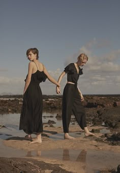 two women in black dresses standing on rocks near the water and holding each other's hands