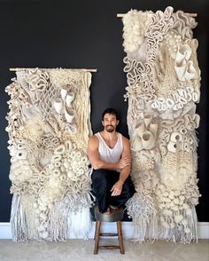 a man sitting on a stool in front of two large wall hangings with tassels