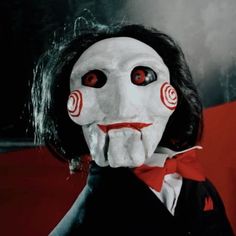 a creepy doll with red eyes and black hair wearing a white mask, holding his hand to his mouth