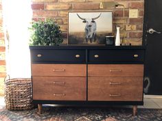 an old dresser with a cow painting on top and some plants in the corner next to it