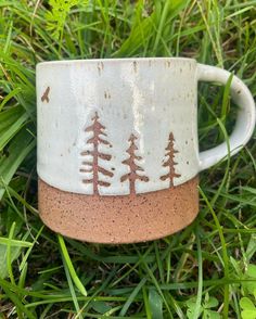 a white and brown coffee cup sitting on top of green grass with trees painted on it