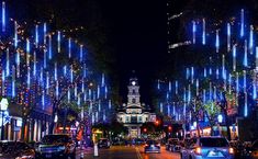 cars driving down a street with christmas lights on the trees and buildings in the background