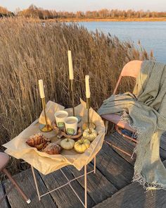 two chairs and a table with food on it near the water in front of some tall grass