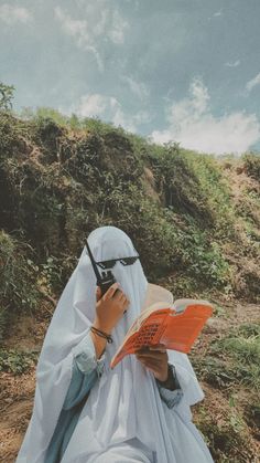 a person sitting down reading a book and talking on a cell phone while wearing a headscarf